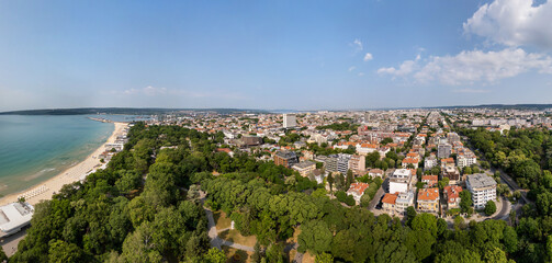 Aerial panoramic view of Varna, Bulgaria, showcasing the urban landscape with red rooftops, green parks, and the Black Sea in the distance under a clear blue sky