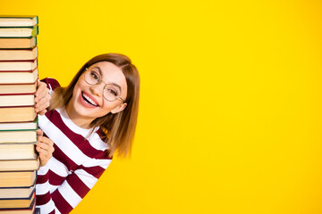 Smiling young woman holding a stack of book against a bright yellow background, expressing cheerful...