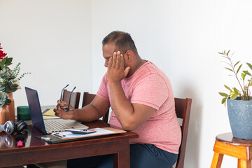 A man in a pink shirt is sitting at a table with a laptop and glasses