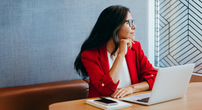 Businesswoman in bright red blazer working on laptop and looking out window