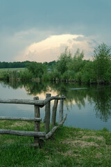 Rustic wooden fence by a calm lake under a dramatic sky, reflecting trees and a bright cloud.