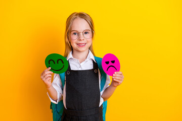 Smiling schoolgirl holding emoticon symbols expressing happy and sad emotions near a yellow background in school uniform