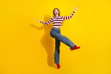 Young joyful female in casual striped pullover and jeans posing playfully against a vibrant yellow...