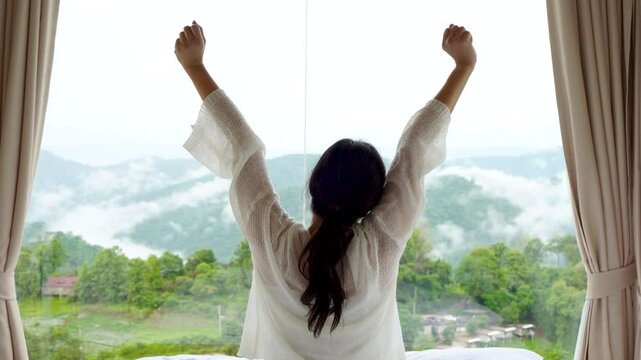 Asian rear woman wake up with her refreshing in morning of holiday with aerial view of white clouds over green trees mountains, she raised two arms. Relax and recharge life by taking a vacation.