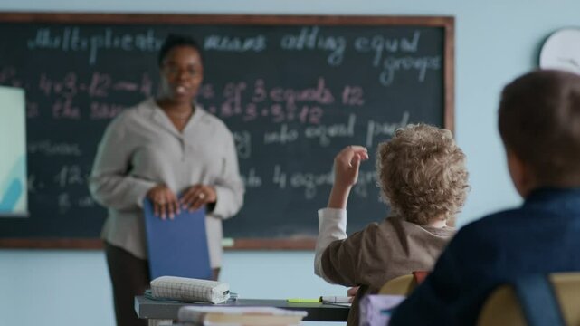 Young curious student raising his hand to ask Black female teacher question on multiplication topic as sitting at desk in front of blackboard - Powered by Adobe