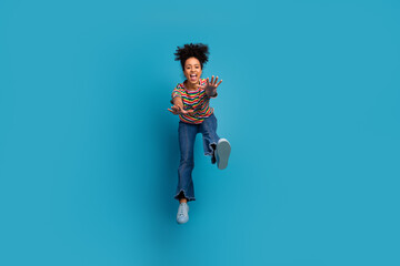 Joyful young girl jumping playfully in colorful striped t-shirt and jeans against vibrant turquoise...