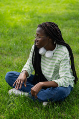 Young african american female student with dreadlocks and glasses is sitting cross-legged on green grass, wearing headphones around her neck and enjoying the peaceful outdoor environment