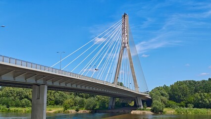 Modern cable-stayed bridge spans over a calm river with lush green trees on both sides. Concept of connectivity, engineering, and urban infrastructure blending with nature