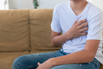 A young man sits on a couch with his hand on his chest