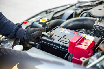 A man wearing gloves loosens a car battery terminal with a wrench under the open hood of a vehicle....