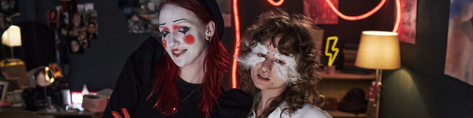 Two Caucasian teenage girls wearing creative makeup standing together in bedroom, one with face...