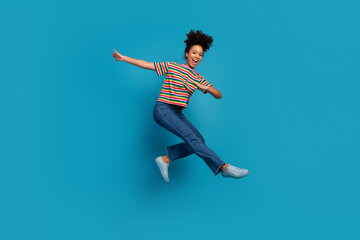 Smiling young woman in casual striped outfit jumps joyfully against a vibrant teal background...