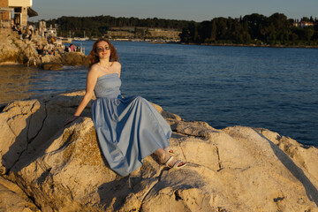 A young woman in a blue dress and sunglasses sits on a rocky shore by the sea at sunset, with buildings and trees in the background.