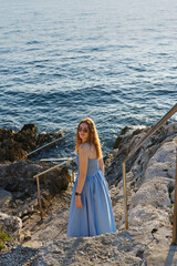 A woman in a flowing blue dress and sunglasses descends rocky steps towards the sparkling blue sea,...