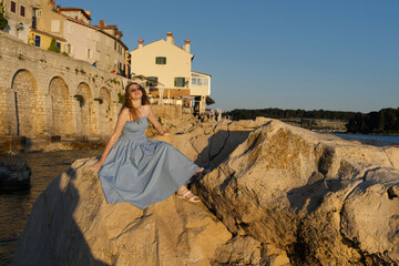 A young woman in a blue dress and sunglasses sits on a sun-drenched rocky shore by the sea,...