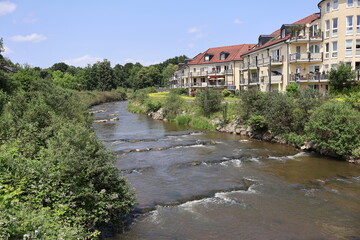 Tranquil stream Mangfall flows through dense greenery next to residential buildings under a sunny...