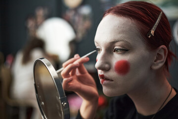 Caucasian teenager girl applying theatrical makeup with face paint and red blush on cheeks using...