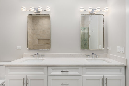 A bathroom detail with white cabinet, marble countertop with chrome faucets, and modern light fixtures above the rectangular mirrors.