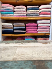 Weathered off-white wooden table with a patina finish in the foreground. In the softly focused background, neatly folded clothes in pastel tones—mainly pink, white, and blue—rest on open closet shelve