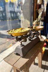 Vintage metal balance scale with two plates, displayed on a wooden bench by a window. One plate holds dried flowers. A soft, nostalgic composition with a quiet view of the street in the background.