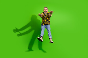 Cheerful middle aged man in a summer shirt jumping joyfully in front of a bright green background,...