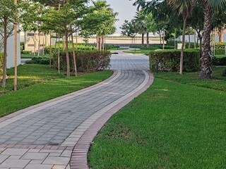 Small Interlock Path Between Landscaping Area.
Green And Lush Landscaping Area With Carpet Grass And Trees Like Palm Or Washingtonia, Some Houses Or Villa's Also Beside Gardens Padestrian Pathway.