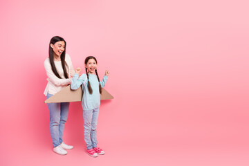 Mother and daughter playing creatively with cardboard airplane prop against a pink background,...