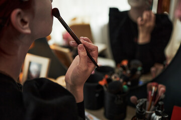 Caucasian teenage girl applying makeup with brush in front of mirror, holding cosmetic tool near face, reflected in glass, demonstrating creative self expression at home