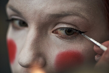 Caucasian teenager girl applying black eyeliner to eyelid with makeup brush, face painted with...