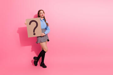 Young female student holding a large cardboard question mark sign against a pink background,...
