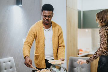 A man in a cozy sweater serves a meal onto a plate in preparation for a casual dinner with peers in...
