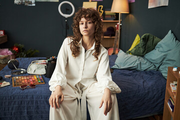 Portrait of Caucasian teenager girl sitting on bed in creative bedroom, looking into camera,...