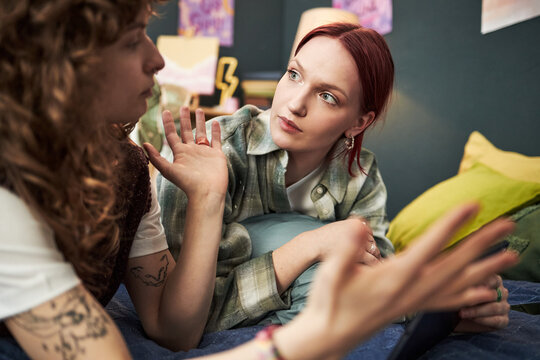 Caucasian teenage girls sitting on bed having animated conversation, one girl gesturing with hands while other girl listening attentively, creative home environment visible in background