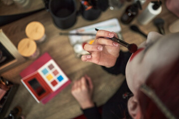 Teenage Caucasian girl applying makeup with brush at home, holding cosmetic palette on wooden...