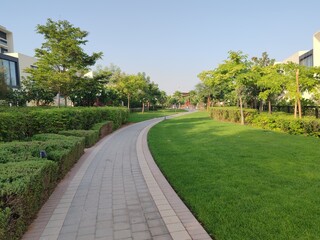 Small Interlock Path Between Landscaping Area.
Green And Lush Landscaping Area With Carpet Grass And Trees Like Palm Or Washingtonia, Some Houses Or Villa's Also Beside Gardens Padestrian Pathway.