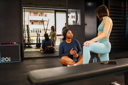 Inside a gym, a personal trainer encourages an overweight woman resting on the floor after exercising, promoting body positivity and healthy lifestyle