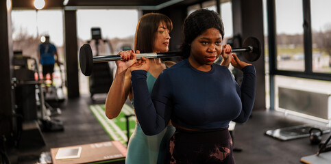 In a modern gym, a determined overweight woman is lifting weights with the assistance of her...
