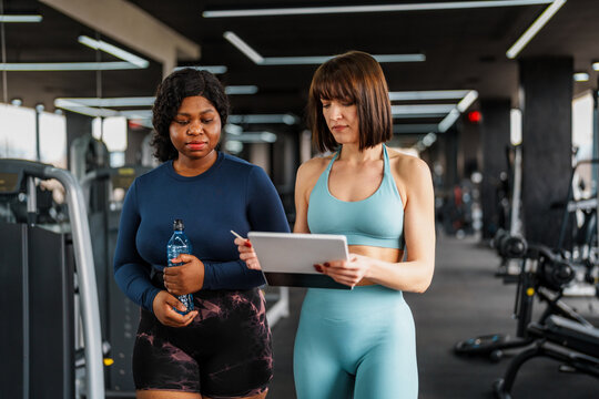 Personal trainer is guiding an overweight woman through a fitness plan on a tablet, providing support and motivation in a gym setting, promoting health and well being