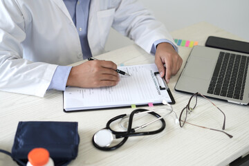 Young professional male doctor is taking notes in journal, physician therapist practitioner filling medical documents with mobile phone or laptop, patient form, illness history, prescription in clinic