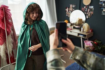 Caucasian teenage girl wearing green cape standing in bedroom smiling while another teenage girl...