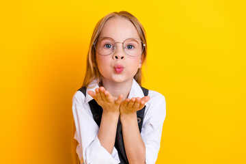 Joyful schoolgirl in glasses sends an affectionate air kiss while posing against a vibrant yellow...