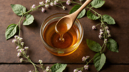 Honey dripping from wooden spoon in glass bowl with organic honey, honey plant acacia flowers and leaves near it on wooden table. Top view.