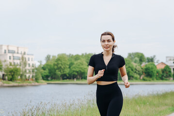 young slim beautiful caucasian woman running on path on river embankment in city park, black...