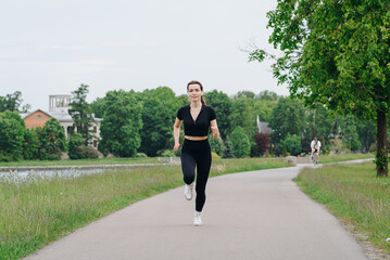 young slim beautiful caucasian woman running on path on river embankment in city park, black...