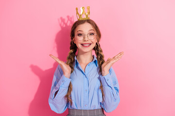 Cheerful young girl with a crown on her head smiling against a pink background expressing joy and...
