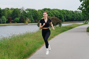 young slim beautiful caucasian woman running on path on river embankment in city park, black...