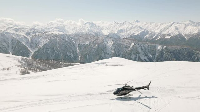 Aerial view of a black helicopter stationed on a snow-covered alpine peak in the Caucasus Mountains of Georgia during clear winter weather. Heliskiing outdoors in alps Europe concept