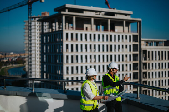 Two architects in safety vests and helmets discuss building plans on a rooftop, with unfinished buildings in the background on a clear day.