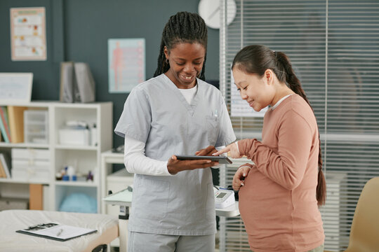 Asian pregnant young adult woman consulting with Black young adult female doctor during prenatal care checkup, both standing in medical office and interacting with digital tablet