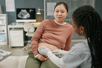 Asian pregnant young adult woman sitting on examination table receiving prenatal checkup from Black...